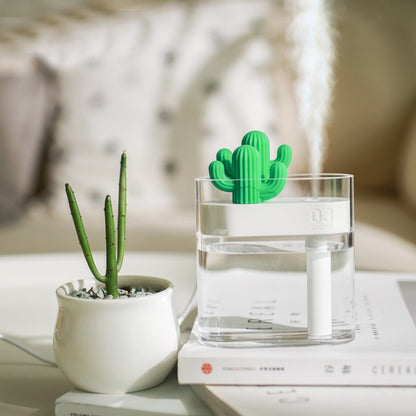 The Valga 160ML Ultrasonic Air Humidifier Clear Cactus Color Light USB Essential Oil Diffuser Car Purifier Aroma Diffusor Anion Mist Maker, designed in a small green cactus shape, is placed in a transparent water-filled container and emits a fine mist. Next to it sits a potted succulent plant. Both items are displayed on a white table against a blurred background decor, offering relief from dry air.