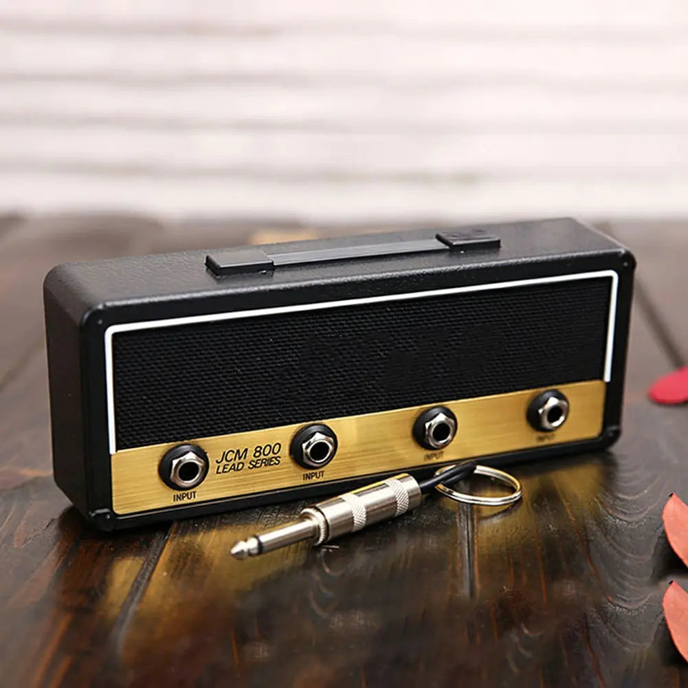 A rectangular, black and gold Valga Wall Mounting Guitar Amp Key Hanger sits on a wooden surface. Embracing the rockstar style, it features three input knobs labeled "INPUT" and "JCM 800 LEAD SERIES." A quarter-inch instrument cable plug keychain is placed in front of this guitar amp key hanger.