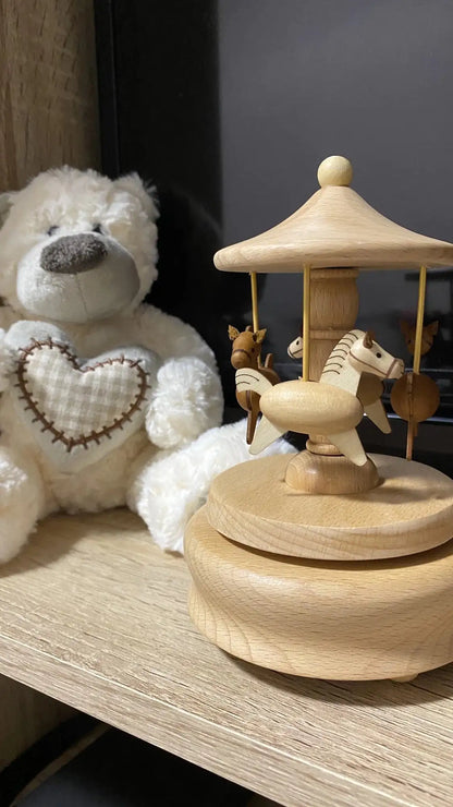 A white teddy bear with a heart patch sits next to the Valga Music Box - Vintage Wood on a shelf. The music box, reminiscent of nostalgic decor, features three wooden horses and has a simple, natural finish. The background is slightly dark, highlighting the bear and the heirloom gift.