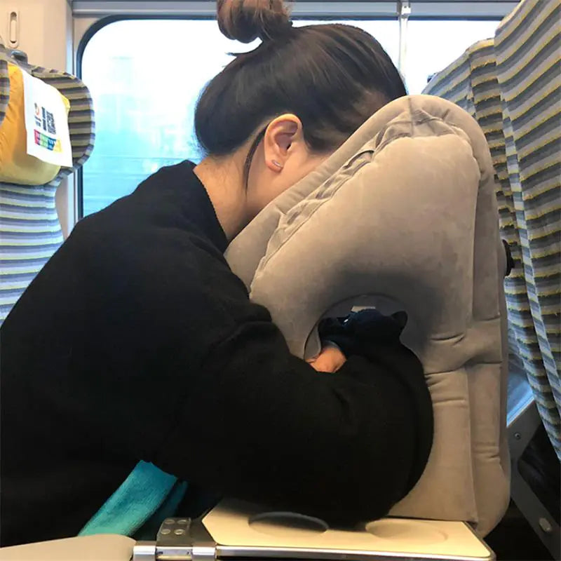 A person with dark hair tied in a bun is seated on a train, leaning forward with their face resting on a grey Valga Inflatable Air Cushion Travel Pillow placed on a foldable table. This compact travel accessory ensures neck pain relief, while the train seats and window are visible in the background.