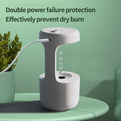 A Valga LED Water Drop Humidifier Diffuser, featuring a sleek white design and whisper-quiet technology, sits on a round, green table against a matching green background. The text "Double power failure protection. Effectively prevent dry burn" is displayed above it. The device is plugged in, with visible bubbles inside it, and a small plant is placed next to the humidifier for added aesthetic appeal.