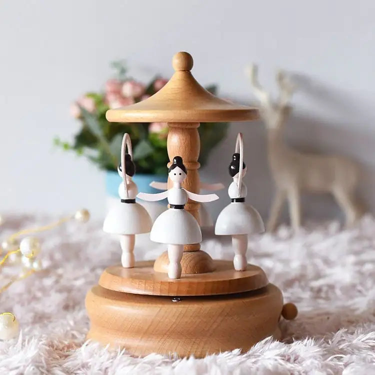 The Valga Music Box - Vintage Wood features three ballet dancers in white dresses gracefully twirling under a pavilion-like canopy. The background is adorned with charming decorative elements, including a potted plant with pink flowers and a small deer statue. This nostalgic piece rests on a fluffy, light-colored surface, making it an ideal heirloom gift.