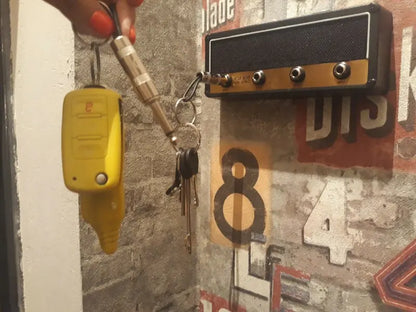 A hand holds a set of keys, including a yellow key fob, up to the Valga Wall Mounting Guitar Amp Key Hanger. The background features decorative graffiti-style numbers and letters on a textured surface, giving the scene an undeniably rockstar vibe.