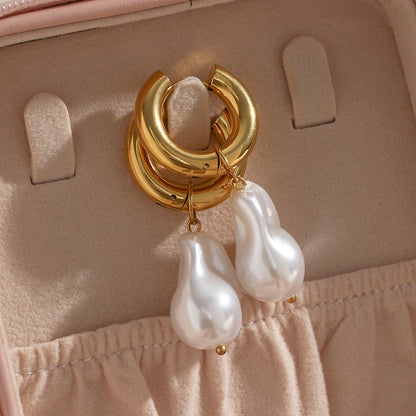 A pair of stunning Valga Water Drop Earrings with large, white baroque pearl charms dangle elegantly against a soft beige fabric background, likely part of an earring holder or jewelry box. The texture and luster of the pearls beautifully contrast with the golden hoops.