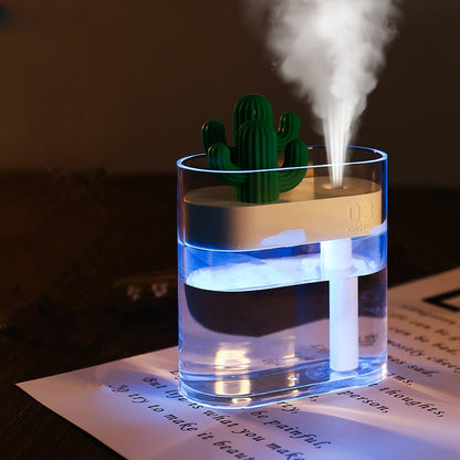 A small, clear 160ML Valga Ultrasonic Air Humidifier with a cactus-shaped top emits a fine mist. The USB Essential Oil Diffuser, illuminated by a blue light, sits on a sheet of paper with printed text, providing relief from dry air.