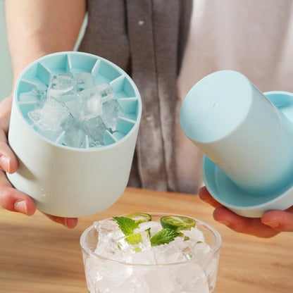 A person is holding the Valga Silicone Cylinder Portable Ice Maker Bucket and its lid over a glass. The mold, filled with ice cubes, is being opened to release the ice into the glass, which also contains lime slices and mint leaves.