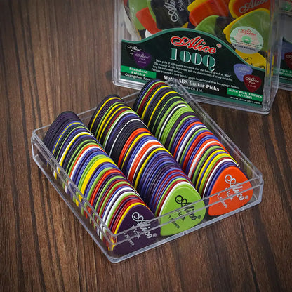 A clear plastic box containing several colorful, durable Valga Guitar Picks arranged neatly in rows. The background shows a wooden surface and parts of a display box with the Valga branding and additional ABS plastic picks visible.