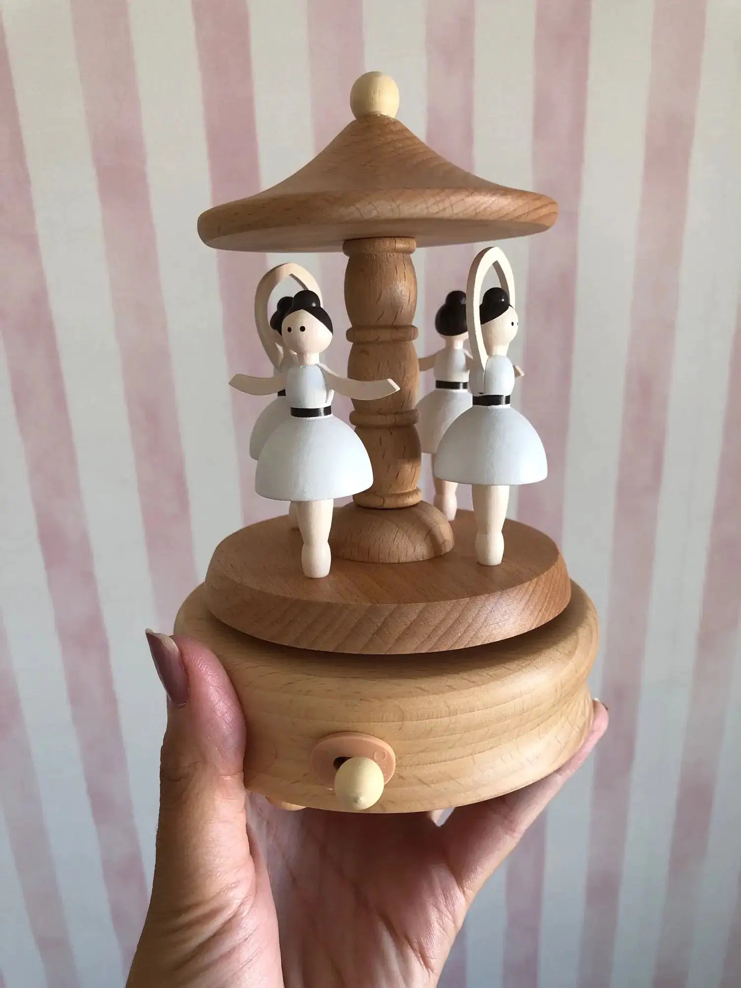 A hand is holding a Valga Music Box - Vintage Wood, a perfect piece of nostalgic decor, featuring three white ballerina figures dancing around a central column under a canopy. The background showcases vertical pink and white stripes, making it an ideal heirloom gift for any cherished collection.