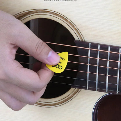 Close-up of a hand holding a durable Valga Guitar Pick in yellow while playing an acoustic guitar. The guitar strings and sound hole are visible, and the background shows the wooden body of the guitar.