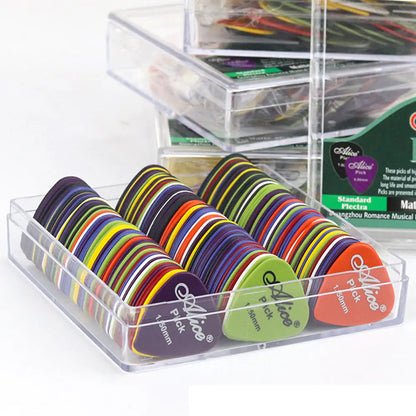 A clear plastic container holds numerous colorful Valga guitar picks arranged in rows. The durable picks, made of ABS plastic, come in vibrant colors such as black, green, red, and yellow. Each pick has "Alice Pick" printed alongside its thickness measurements. Additional containers are visible in the background.