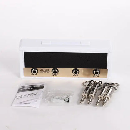 A Wall Mounting Guitar Amp Key Hanger by Valga, labeled with "JCM 800 Lead Series" above four simulated input jacks, rests on a white surface, exuding rockstar style. Three guitar cable keychains, screws, and an instruction manual are placed in front of the white-cased hanger with a black front panel.