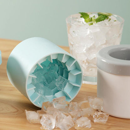 A Valga Silicone Cylinder Portable Ice Maker Bucket in light blue, with ice cubes spilling out, is placed on a wooden surface. Next to it is a glass filled with crushed ice and garnished with mint leaves. Another part of the ice tray mold is visible in the background.