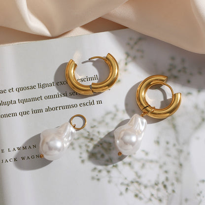 Close-up of Valga's Water Drop Earrings: one with a dangling baroque pearl charm and the other detached. The gold hoop earrings rest on an open book featuring Latin text, partially draped with light-colored fabric in the background.