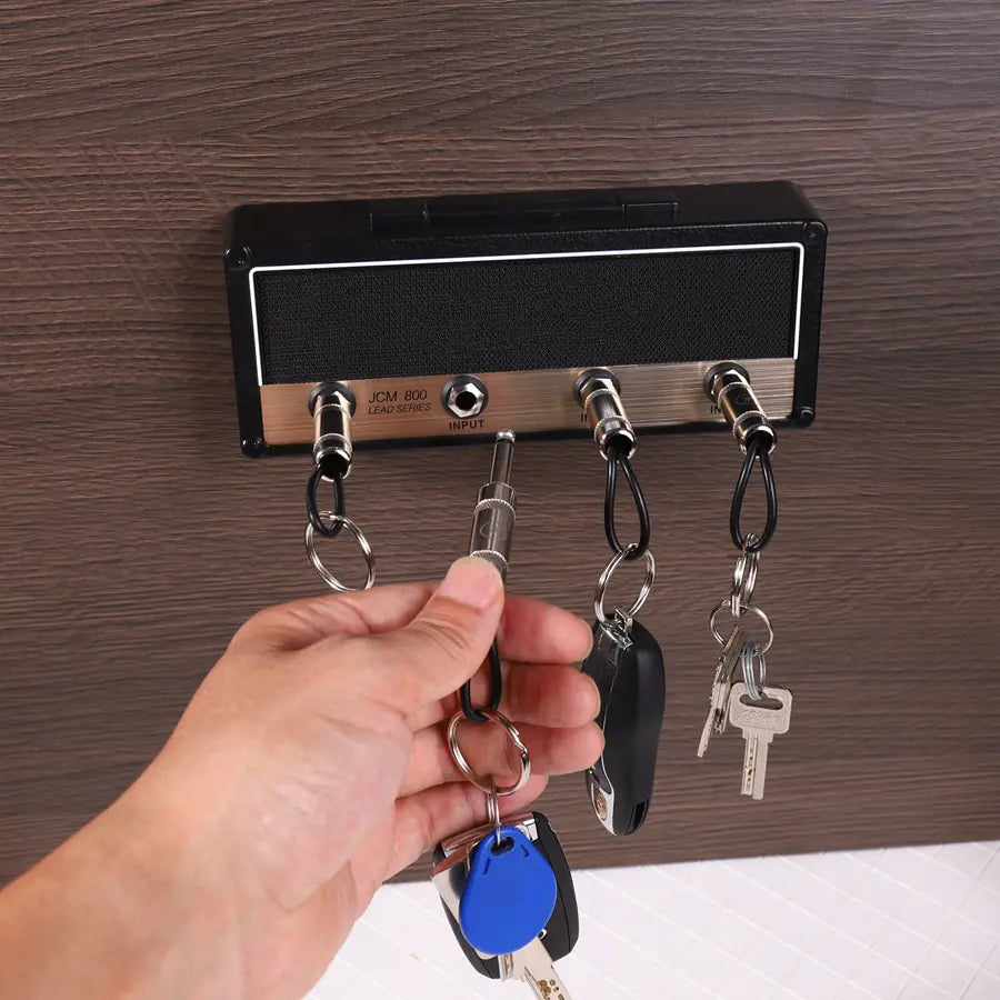 Valga's Wall Mounting Guitar Amp Key Hanger is crafted to resemble the front panel of a guitar amplifier. It features four input jacks, each holding guitar plug keychains with various keys. A hand is shown plugging in a keychain with a blue key fob into one of the jacks, perfectly capturing that rockstar vibe.