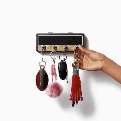 A hand holds keys attached to a colorful tassel keychain next to guitar plug keychains hanging from hooks on a rectangular Wall Mounting Guitar Amp Key Hanger by Valga. The black holder with a gold plate and five hooks radiates rockstar style, adorned with other keychains like pom-poms and small pouches.