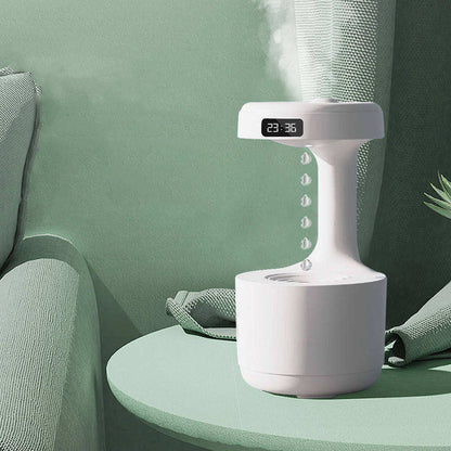 A white LED Water Drop Humidifier Diffuser with a minimalist design stands on a green round table. This Valga product features an LED clock display and a unique structure where it appears as if water drops are suspended mid-air between the base and the light source. It is placed beside a green armchair.