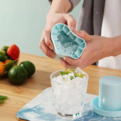 A person is seen squeezing a Valga Silicone Cylinder Portable Ice Maker Bucket to release ice cubes into a glass filled with ice and lime slices. The glass sits on a blue and white patterned mat on a wooden surface, with assorted fruits like limes and a pomegranate in the background.