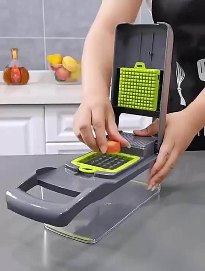 In a modern kitchen, a person expertly uses the Valga Vegetable Chopper Kitchen with a green grid blade to slice through a piece of carrot. On the gray counter, surrounded by white cabinets and a glass pot holding fruits, this scene epitomizes efficient meal preparation.