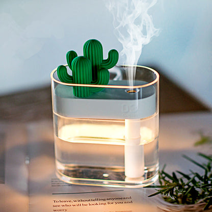 A small Valga 160ML Ultrasonic Air Humidifier Clear Cactus Color Light USB Essential Oil Diffuser sits on a wooden surface next to a leafy green plant, providing dry air relief with its fine mist. The humidifier, containing water and emitting a soft, ambient light, features a charming green cactus-shaped ornament on top.