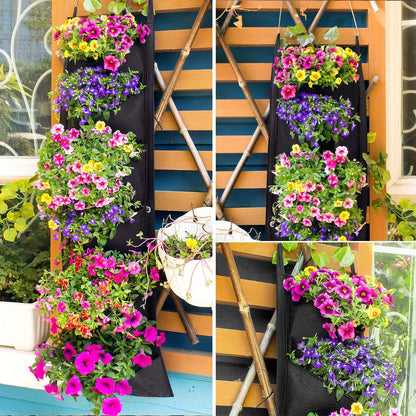 A wooden fence features two Valga Vertical Hanging Garden Flower Pots in black, filled with vibrant flowering plants. The flowers are a mix of pink, purple, yellow, and white blooms. Potted plants and climbing vines add to the lush greenery around the fence.