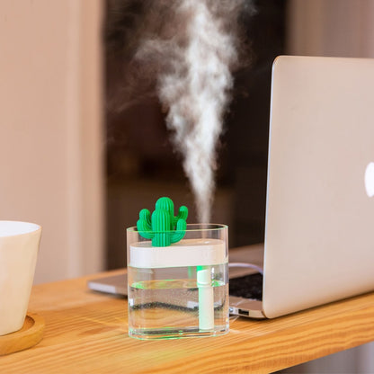 A Valga 160ML Ultrasonic Air Humidifier Clear Cactus Color Light USB Essential Oil Diffuser emits a fine mist from a transparent glass of water, positioned on a wooden desk next to a silver laptop. Also seen on the desk is a white bowl on a coaster, providing relief from dry air. The background is slightly blurred.