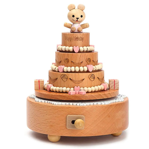 The Valga Music Box - Vintage Wood, styled like a three-tiered birthday cake adorned with small heart and bead decorations, serves as nostalgic decor. At the top, a smiling bear figurine is featured, while small red and white presents surround the base. This charming heirloom gift includes a winding key for operation.